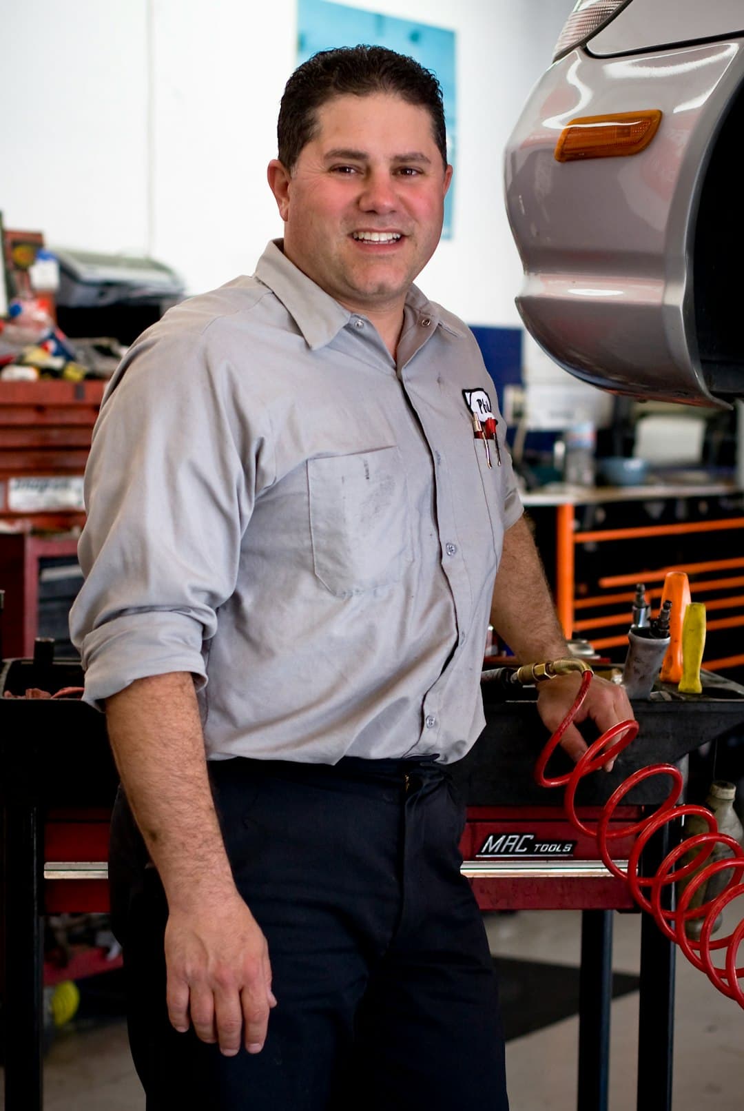 A man in a work shirt smiling beside a car on a lift
